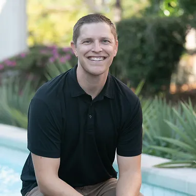 A man in a black polo shirt is sitting beside a pool, smiling at the camera. The background includes greenery and blurred foliage, suggesting an outdoor setting.