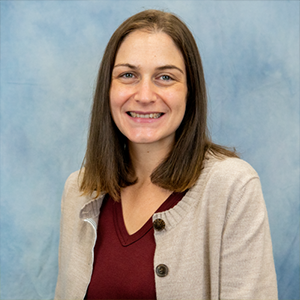 A woman with shoulder-length brown hair smiles at the camera. She is wearing a beige cardigan over a maroon top. The background is a soft blue gradient.